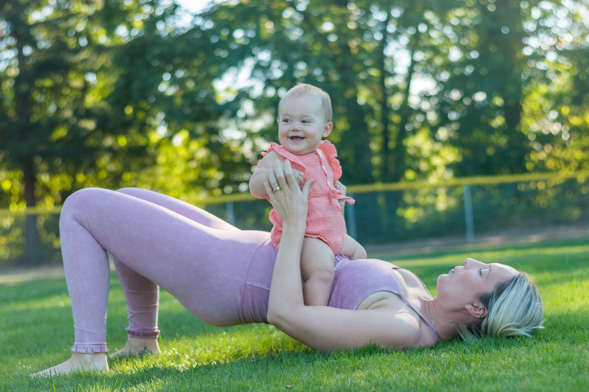 A mother in activewear exercises with her baby in a sunny park.
